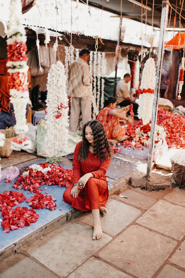 A woman in red sits surrounded by colorful flowers at a lively market, embracing cultural beauty.